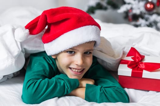 Child boy in green xmas pajamas holds a Christmas gift in his hands while sitting in bed. Generation Alpha and Gen Alpha children.