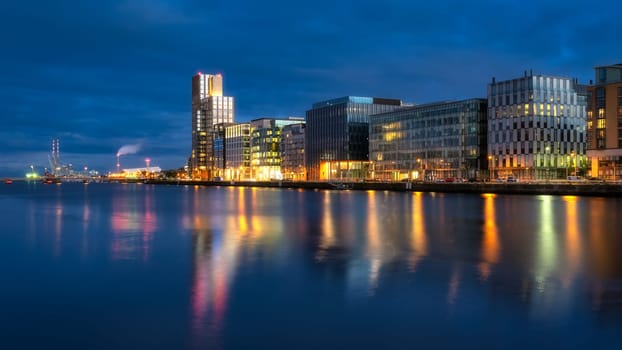 Modern offices and apartment buildings on Sir John Rogerson Quay and Poolbeg Powerstation chimneys, blurred Liffey River at dusk, Dublin, Ireland