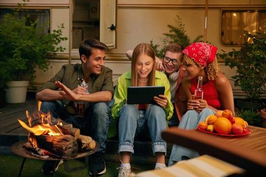 Happy friends spending evening nearby bonfire and campsite. Four millennial people watching video using mobile tablet smiling and laughing. Carefree weekend time on nature concept
