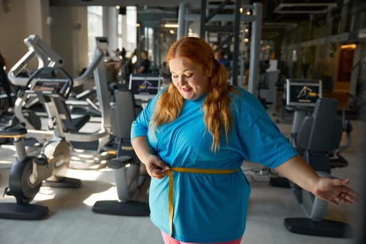Happy satisfied overweight woman seeing first good result on measuring tape after her hard training workout challenge at gym for slimming