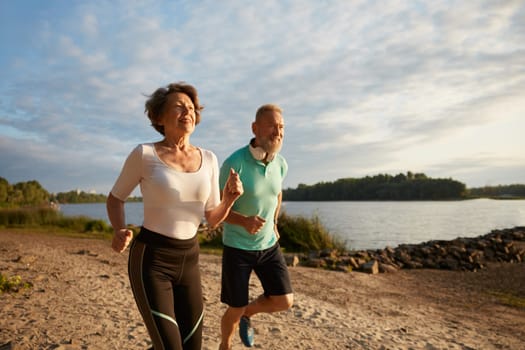 Happy elderly couple running on beach by sea enjoying jogging workout in early morning. Active retirement and healthcare physical exercises outdoors