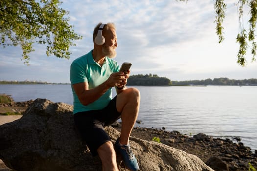Senior man taking break for rest on stone by river after run training in morning. Active elderly sportsman wearing headset listening to favorite music tracks on smartphone for motivation