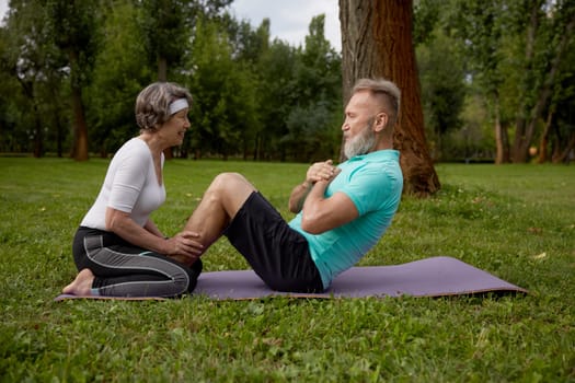 Happy married couple of retirees doing training workout in park. Elderly wife helping senior husband doing abdominal crunch. Sports leisure activity for pensioner