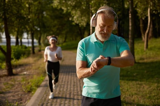Senior man running in park during early morning. Elderly sportsman wearing headset looking at wristwatch fitness tracker caring for his health