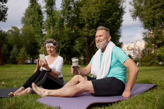 Senior couple sitting on fitness mats rest after yoga training outdoors in park. Elderly woman using smartphone, mature man drinking water. Sports weekend and free time on retirement