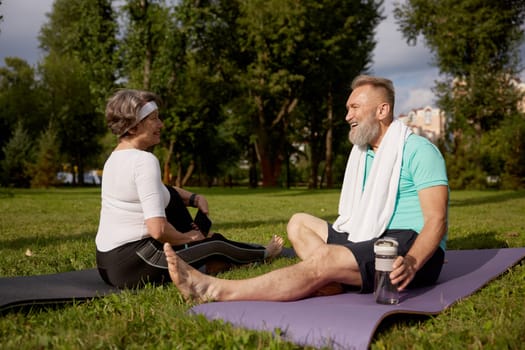 Senior couple sitting on fitness mats rest after yoga training outdoors in city park. Elderly woman using smartphone, mature man drinking water. Sports weekend and free time on retirement