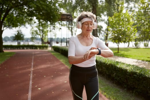 Senior woman running in park during early morning. Elderly sportswoman wearing headset looking at wristwatch fitness tracker caring for his health