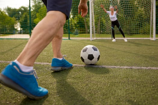 Happy retired people playing football together outdoors at city court. Closeup view on male legs kicking ball and woman in catching position standing at soccer gates