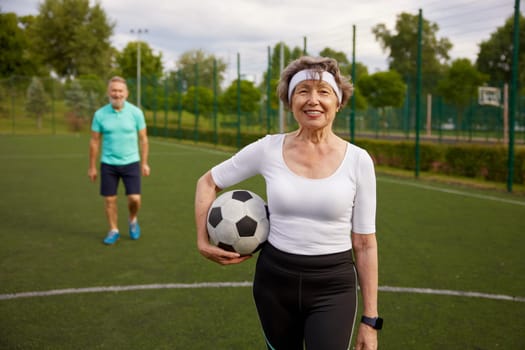 Happy elderly couple with soccer ball on sportive stadium in city park. Healthy habits of retired people