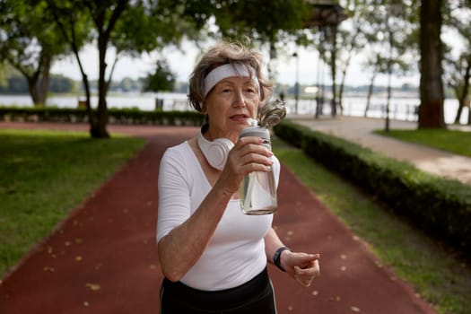 Elderly woman drinking water taking short break during running exercise on stadium field at natural park. Sport hobby activity on retirement