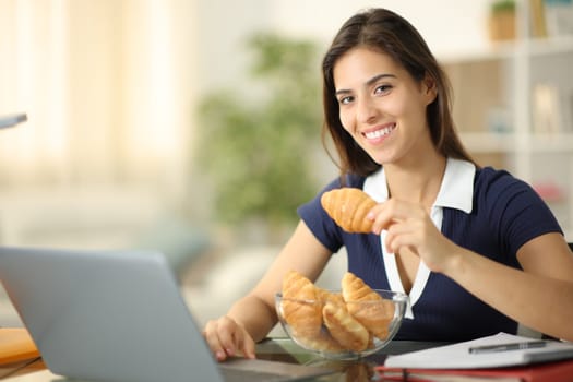 Happy student e-learning eating croissants looking at camera at home