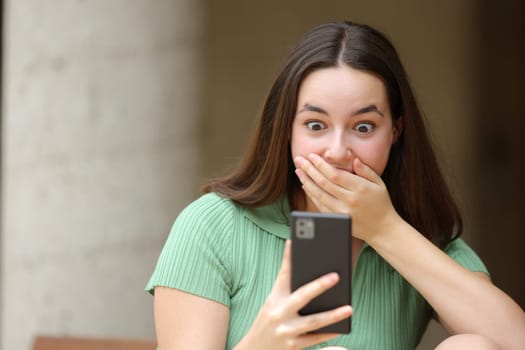 Front view portrait of a surprised woman checking cell phone news in the street