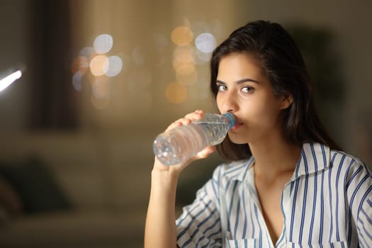 Woman looking at camera drinking bottled water in the night at home