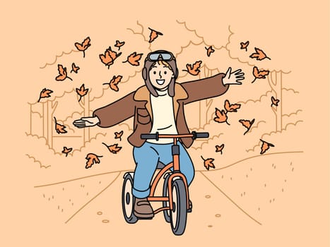 Happy child rides bicycle through autumn park and spreads arms to sides enjoying good weather and falling leaves. Cheerful boy cyclist takes airplane pose riding bicycle along autumn streets