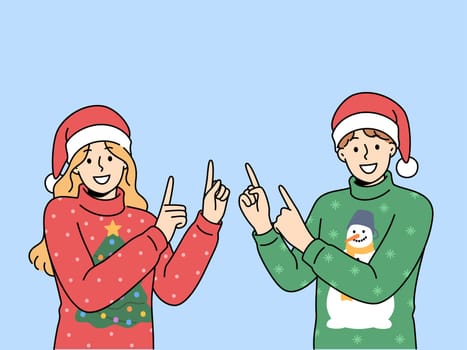 Man and woman in christmas sweaters point fingers upward to visit new year sale. Happy guy and girl in santa hats smiling looking at screen inviting you to christmas party or xmas festival
