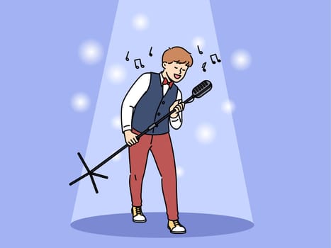 Talented boy singer holds professional microphone on tripod and performs at concert standing in light of spotlight. Happy teenage child singer dreams of becoming star and touring world