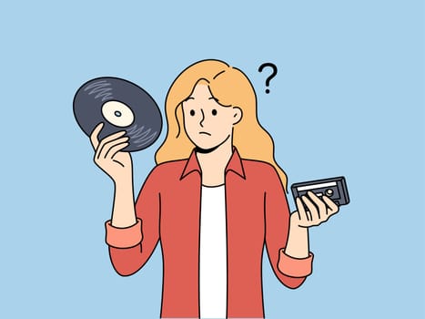 Girl with vinyl record and tape cassette looks confusedly at old-fashioned storage media with music. Woman feels confused and doesn know how to listen to music stored on vintage storage devices.