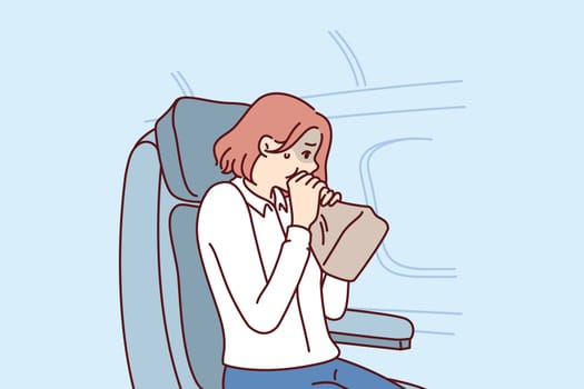 Woman passenger of airplane suffers from aerophobia and breathes into paper bag, sitting in airliner near porthole. Girl client of airline is stressed due to aerophobia or getting into turbulence zone