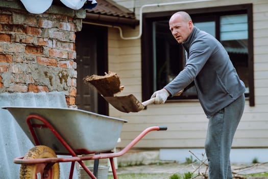 Middle aged man with bald head in gray tracksuit throwing earth into wheelbarrow using shovel while standing near house. Human in gloves collecting land and cleaning ground on countryside yard.