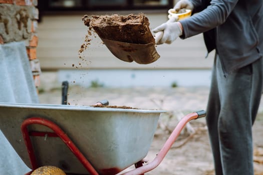 Unrecognizable human in gloves standing near countryside house, holding spade and throw earth into wheelbarrow. Gardener in comfortable tracksuit removing land from lot, working with soil.