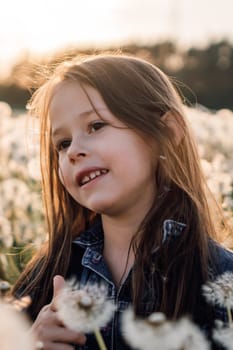 Portrait of charming girl with brown hair standing in meadow full of white dandelions on summer day. Female kid with dark eyes in blue denim jacket looking into distance with soft glance. Vertical.