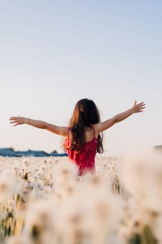 Rear view of little girl in pink light clothes standing in white dandelions meadow with arms wide open. Child with brown long hair enjoying sunny day in nature surrounded by blooming. Vertical.