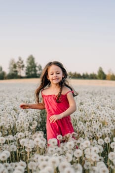 Loveable little girl with dark long hair running across field full of white dandelions and smiling happily. Female child in pink jumpsuit spending time outdoors and walking in flowers. Vertical.