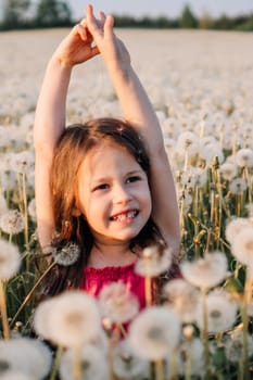 Loveable female child with bright smile standing surrounded by white fluffy dandelions in field and stretching arms up. Happy girl with brown hair in pink summer clothes enjoying sunny day. Vertical.