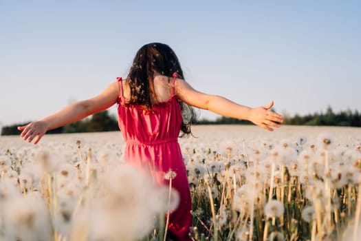 Rear view of female child in pink summer jumpsuit walking in field full of white dandelions with arms wide open. Little girl with dark hair looking down at blossoms and enjoying vacation.