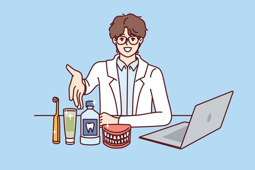 Man dentist pulls hand to screen, sitting at table with laptop and supplies for dental care. Young doctor dentist recommends purchasing toothbrush and toothpaste to maintain hygiene