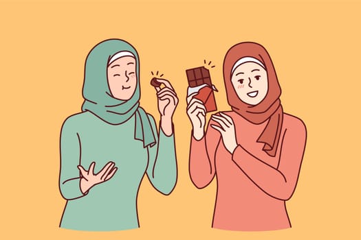 Two arab women eat chocolate and rejoice in rush of hormone of happiness, dressed in islamic scarves and clothes. Girls recommend trying chocolate made from natural cocoa beans and organic cream