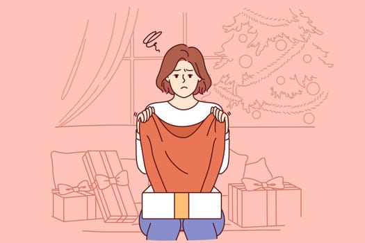 Sad woman upset by christmas gift, sitting on sofa among giftboxes near new year tree. Girl experienced stress after receiving unwanted gift or poor-quality item during christmas holidays