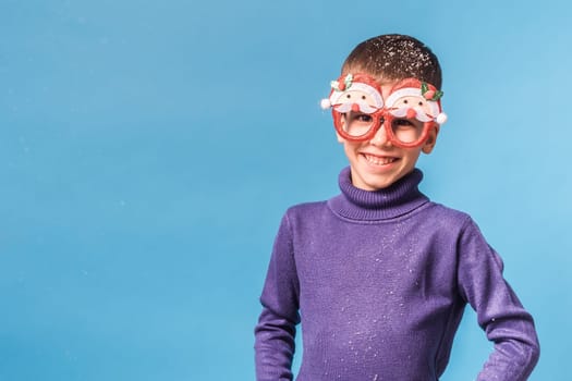 Cute little child boy in party Christmas Santa Claus glasses on blue background. Kid with snow on head copy space and mockup.