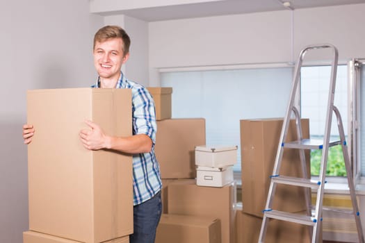 Moving to a new apartment. Cheerful young man holding a cardboard boxes and smiling at camera while other carton boxes laying on background