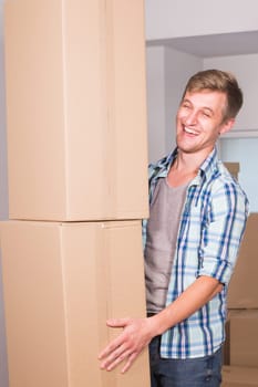 Moving to a new apartment. Cheerful young man holding a cardboard boxes and smiling at camera while other carton boxes laying on background