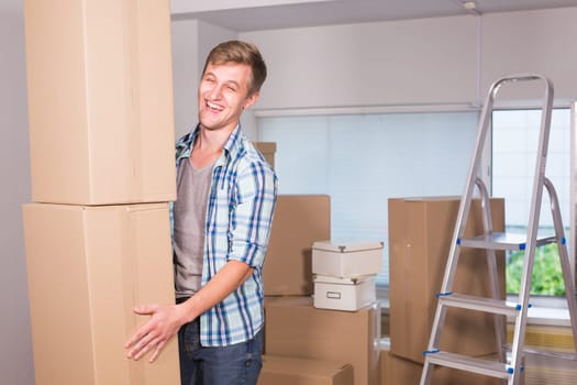 Moving to a new apartment. Cheerful young man holding a cardboard boxes and smiling at camera while other carton boxes laying on background