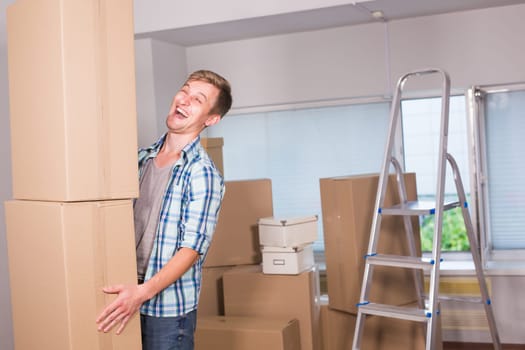 Moving to a new apartment. Cheerful young man holding a cardboard boxes and smiling at camera while other carton boxes laying on background