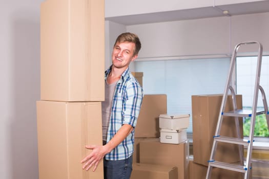 Moving to a new apartment. Cheerful young man holding a cardboard boxes and smiling at camera while other carton boxes laying on background