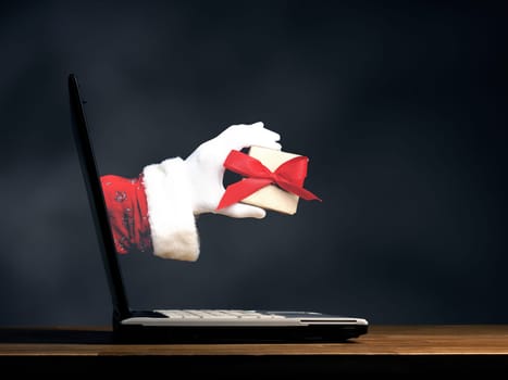 Hand of Santa is holding present with a red bow. Open laptop on a wooden table, Christmas shopping or internet shopping concept