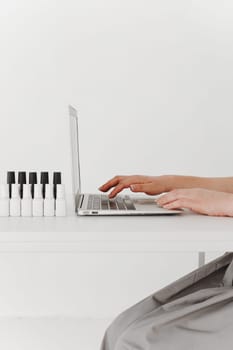 Woman working on the laptop keyboard, jars of nail polish on table, side view, close up on hands, white background