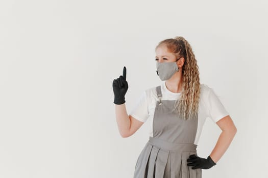 Woman beauty industry nail master in grey uniform and face mask and black gloves shows a sign with finger attention on white background