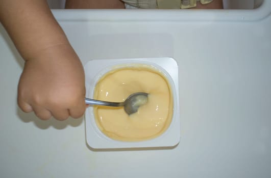 Baby boy eating by himself with spoon. Overhead view of custard bowl