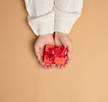 Two female hands holding a red box tied with a ribbon on a brown background, top view