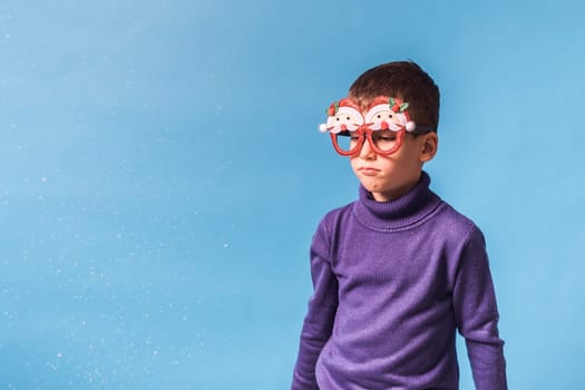 Cute little child boy in party Christmas Santa Claus glasses on blue background. Kid with snow on head copy space and mockup.