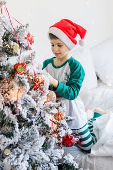 Child decorates Christmas tree. The boy is holding a Christmas toy. Morning before christmas at home. Generation alpha and gen alpha children