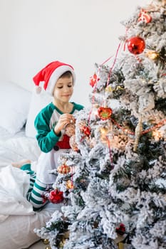 Child decorates Christmas tree. The boy is holding a Christmas toy. Morning before christmas at home. Generation alpha and gen alpha children