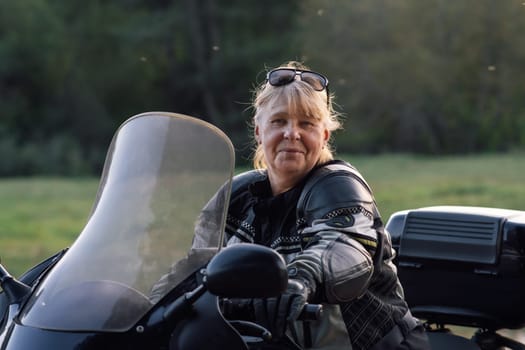 Happy smiling blonde middle-aged motorcyclist in leather jacket, black gloves is sitting on her bike on autumn day. Hobby . Healthy . Emotional relief. Excellent physical shape.Tourism and travel.