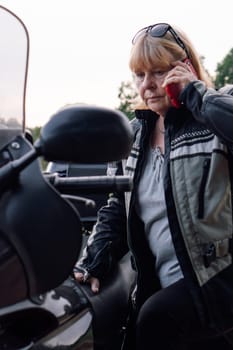 Vertical portrait of mature blonde biker who is talking on mobile phone while sitting on her motorcycle. Communication in travel. Hobby - motorcycle tourism. Female biker. Tourism . Strong women.