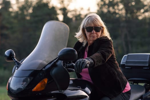 Stylish blonde motorcycle tourist, super mom, grandmother in black gloves and black glasses, sitting on her bike. Hobby motorcycle tourism. Healthy lifestyle. Excellent physical shape.Tourism travel.