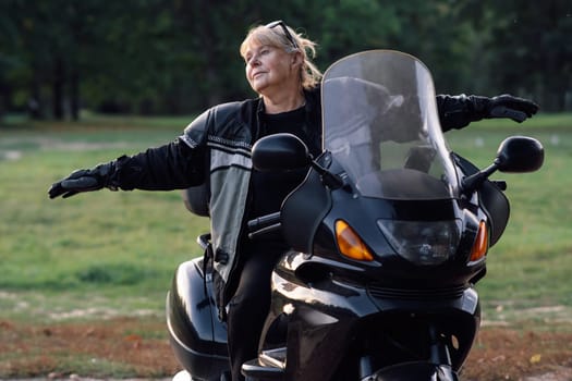 Towards adventures. Happy mature blonde motorcyclist has her arms outstretched, sitting on her black bike. Hobby motorcycle tourism. Emotional relief. Excellent physical shape. Tourism and travel.